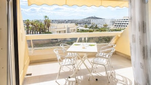 Een balkon met een tafel en stoelen, een geel zonnescherm en uitzicht op gebouwen en palmbomen.