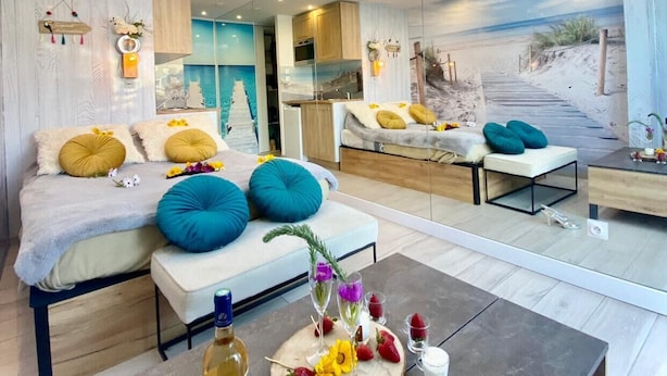 Een modern interieur met een groot dakraam, een gezellige zithoek met kussens, een eettafel met een fles en glazen, en een muurschildering met een strandthema.