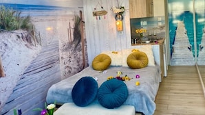 Een moderne slaapkamer met een strandmuurschildering, een bed met twee grote kussens, een nachtkastje met drankjes en uitzicht op een zwembad door een glazen wand.