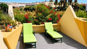 Terraza en la azotea con dos sillones de jardín verdes, un macetero con flores rojas y vistas a palmeras y edificios.