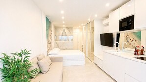 A modern hotel room with a sofa, a small bed, a kitchen area with a microwave, and a large window. - Naturiste Cap d'Agde Héliopolis AB 30 (Agde)