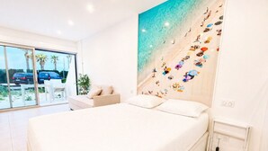 A bedroom with a large bed, a sofa, a window with a view of parked cars, and a wall with a beach-themed mural. - Naturiste Cap d'Agde Héliopolis AB 30 (Agde)