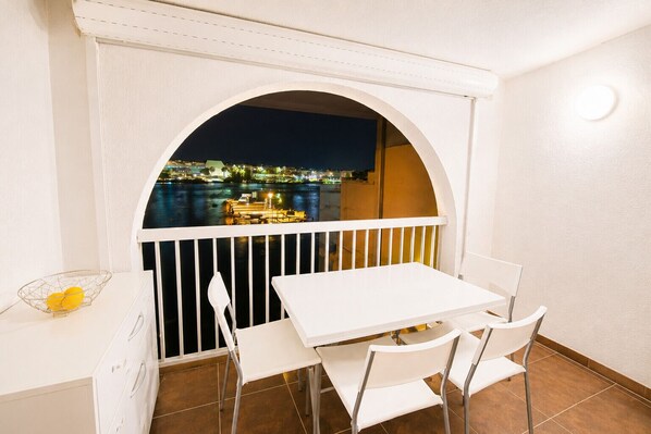 Uma varanda com mesa e cadeiras brancas, corrimão branco e vista para a paisagem urbana à noite.