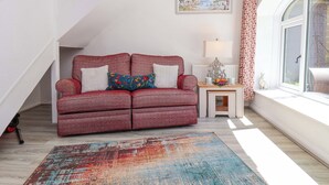 Ein gemütliches Wohnzimmer mit einem roten Sofa, einem bunten Teppich, einem kleinen Tisch mit einer Lampe und einem Fenster mit Blick ins Freie.
