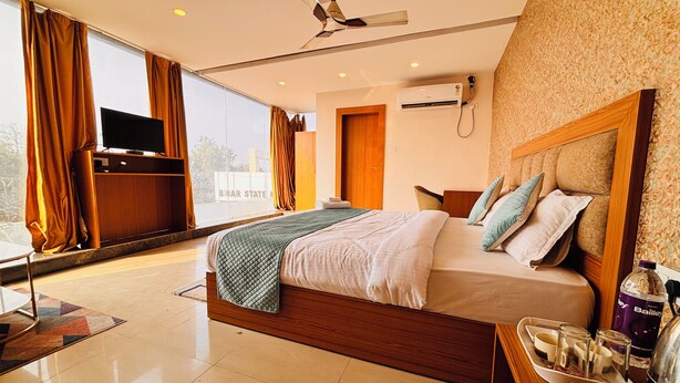 A modern hotel room with a large bed, a flat-screen TV, a ceiling fan, and a view of the city. - Aqoss Hotel (Patna)