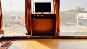 A room with a wooden TV stand and curtains, a tiled floor, and a view of a busy street and buildings. - Aqoss Hotel (Patna)