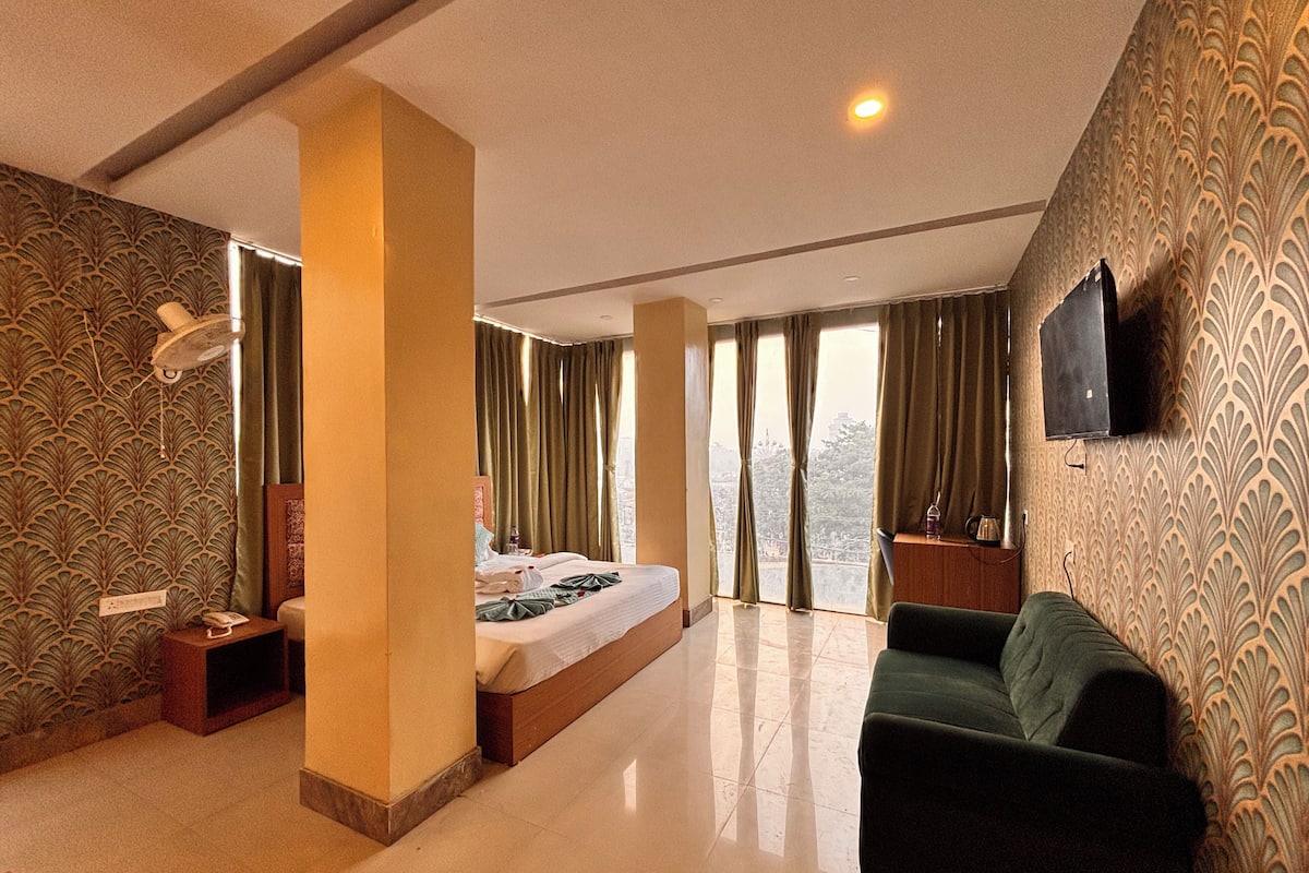 A hotel room with a bed, a green sofa, a TV mounted on the wall, and a large window with curtains.