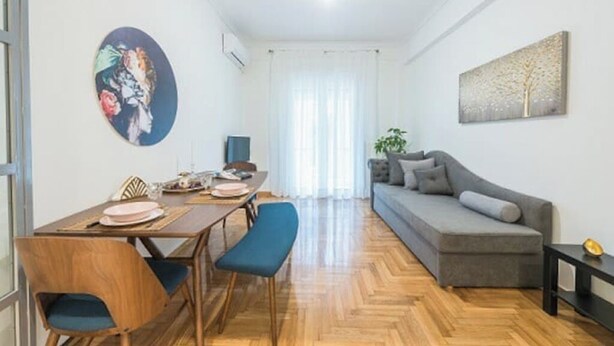 A modern living room with a grey sofa, wooden dining table, and blue chairs. - Cozy home in Athens (Nea Smirni)