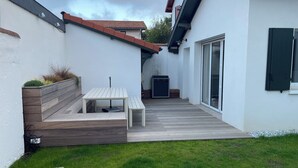 A modern outdoor patio with a wooden table and benches, surrounded by a white wall and a grassy lawn.