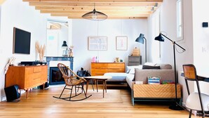 A spacious living room with wooden floors, a sofa, a coffee table, and a dining area. A surfboard is mounted on the wall, and there is a staircase leading to an upper level.