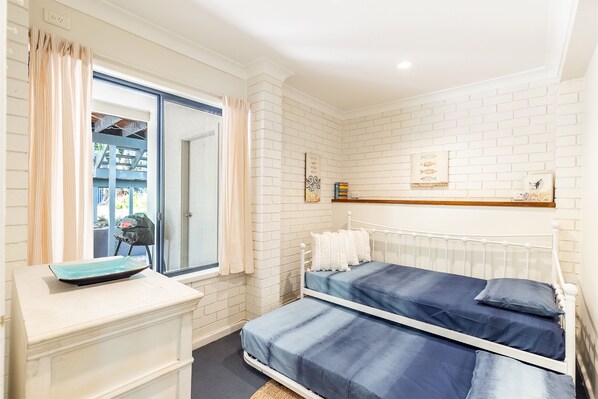 A bedroom with a white brick wall, a bed with blue bedding, a white dresser, and a window with curtains.