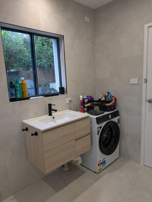 A compact laundry room with a washing machine, a sink, and a window overlooking a backyard.