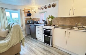 A compact kitchen with a built-in oven, a sink, and a microwave. There is a window with curtains, a shelf with dishes, and a potted plant on the counter.