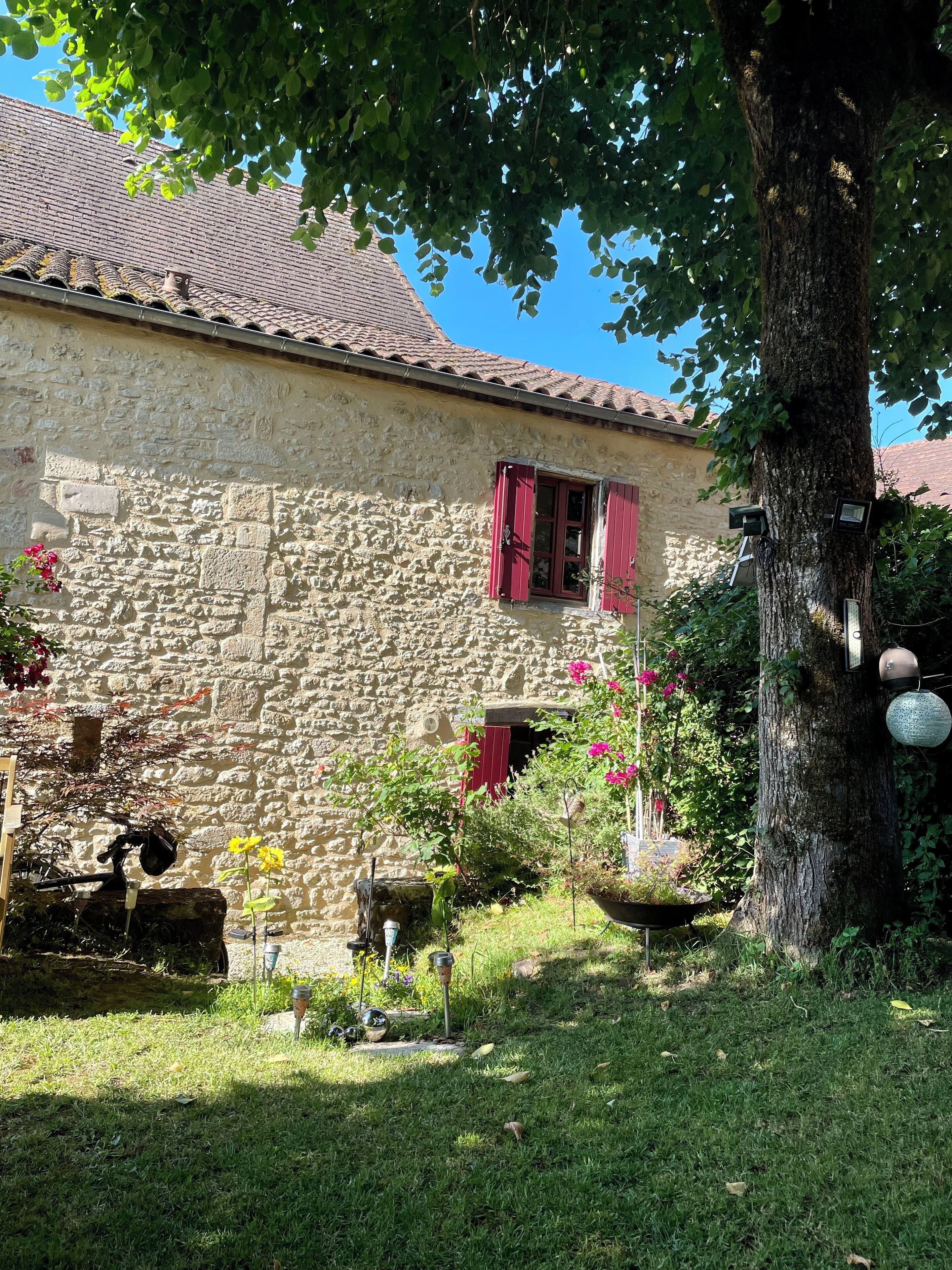 Un bâtiment en pierre avec un toit en tuiles, un arbre au premier plan et un petit jardin agrémenté de fleurs et d’un abreuvoir pour oiseaux.