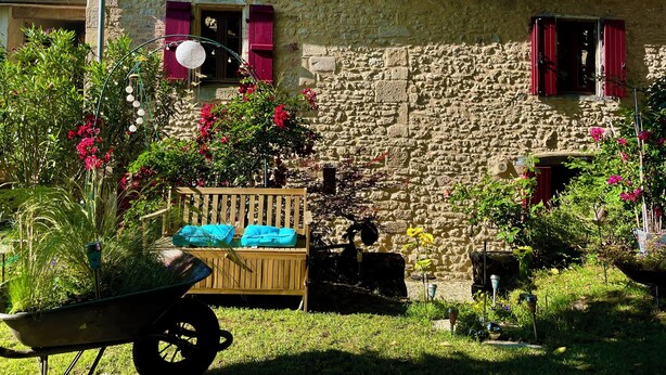 Una casa de piedra con contraventanas rojas, un banco de madera y un carretón en el jardín.