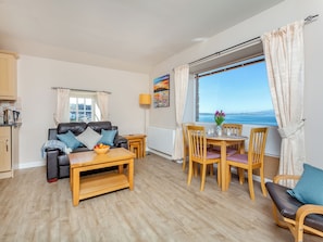 Un salon chaleureux avec un canapé, une table basse et un coin repas. Une fenêtre offre une vue sur la mer.