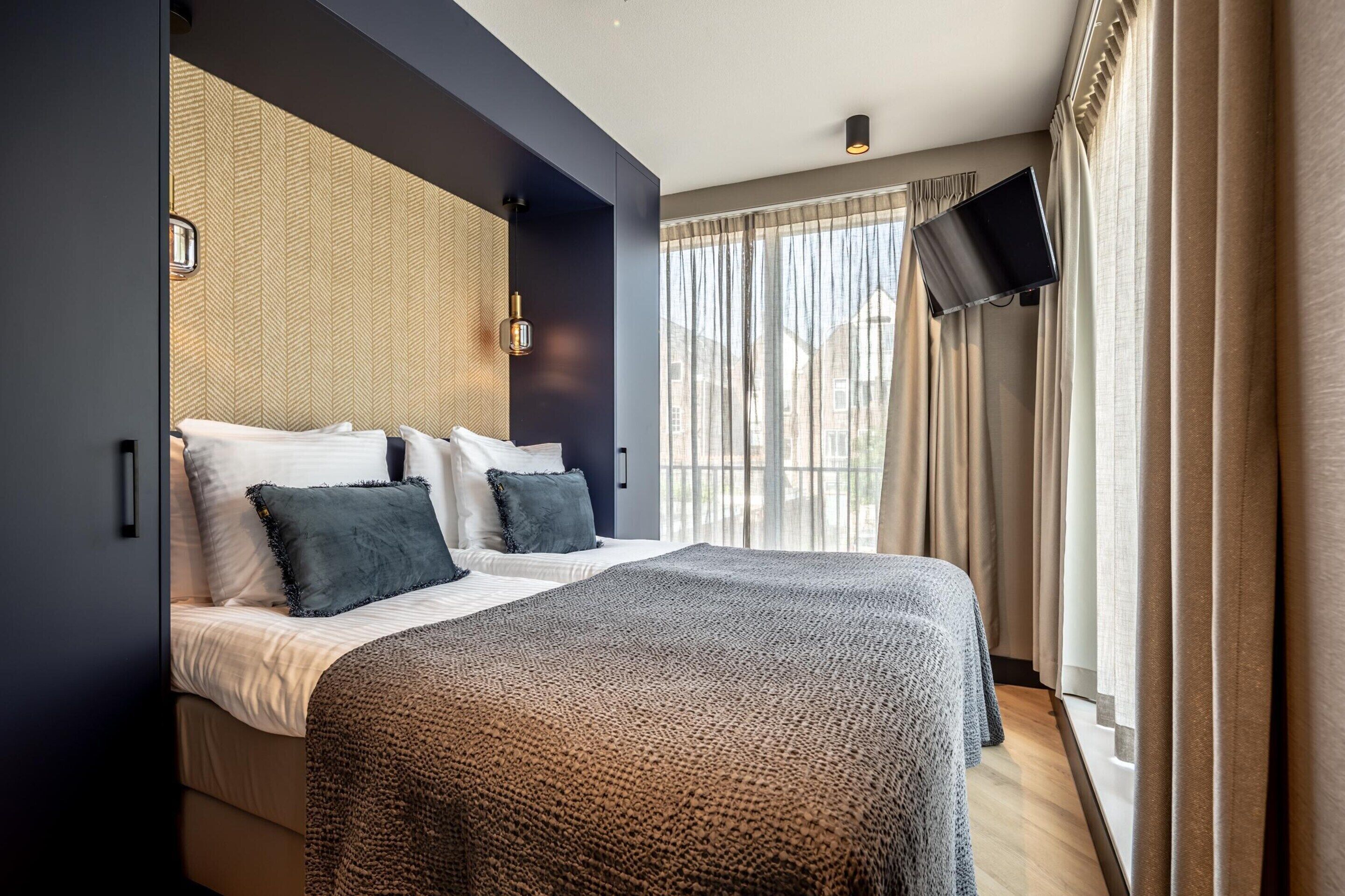 Quarto de hotel moderno com uma cama grande, televisão de tela plana na parede e vista da cidade através de amplas janelas.