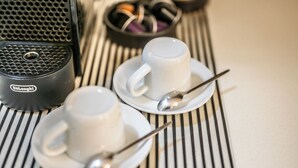 Two white cups on saucers with spoons, a black coffee maker, and a bowl of assorted chocolates on a striped table. - Emilia Studios 5 (Modena)
