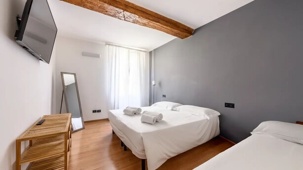 A hotel room with a bed, a bedside table, a TV mounted on the wall, and a large window with curtains. - Emilia Studios 5 (Modena)