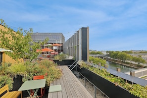 A rooftop terrace with outdoor seating, a view of a river, and a modern building in the background. - All Suites Hôtel | Paris Pleyel – L’île Saint Denis (L'Île-Saint-Denis)