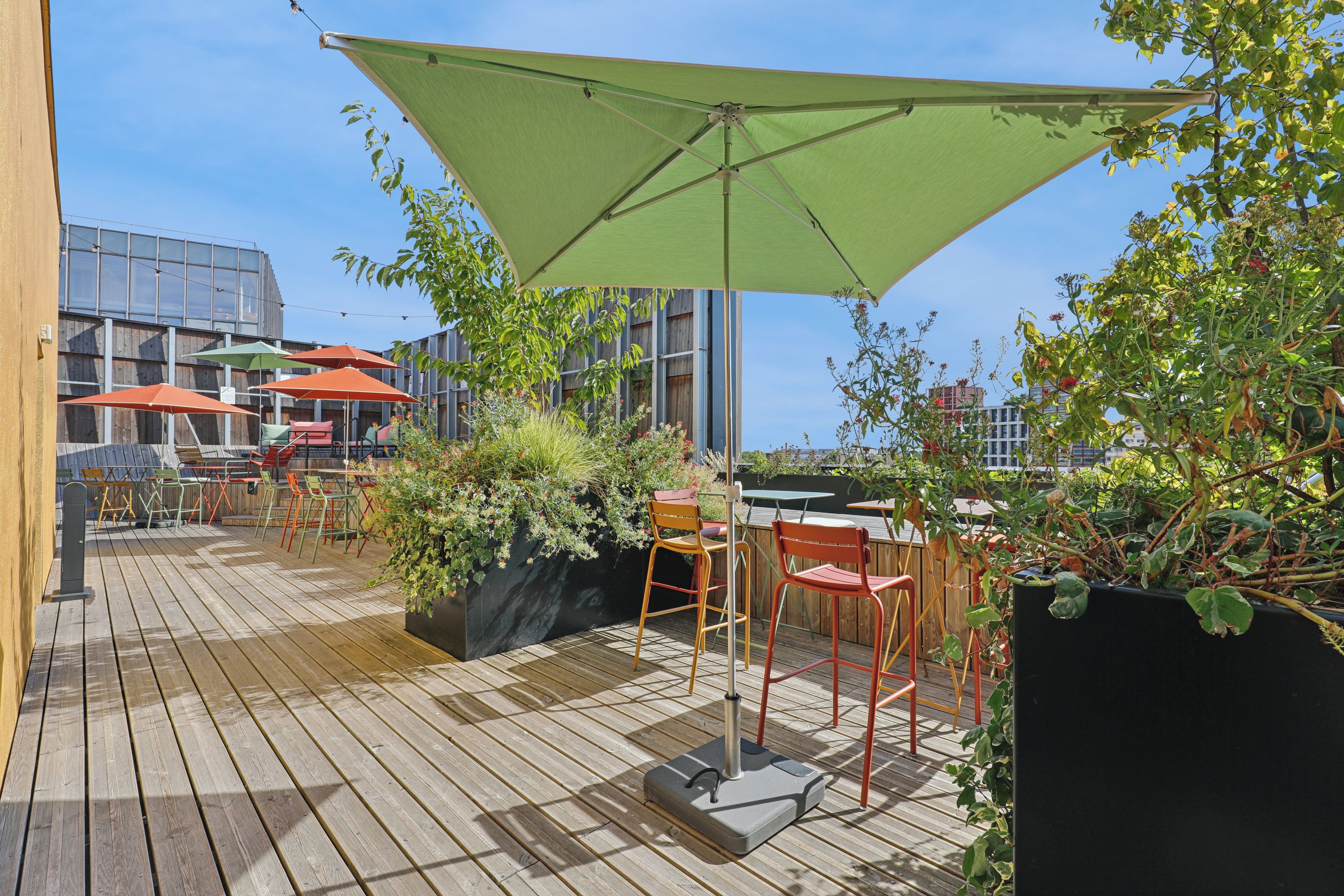 Un espace extérieur aménagé en terrasse, avec un sol en bois, plusieurs tables et chaises, et un grand parasol vert offrant de l’ombre.