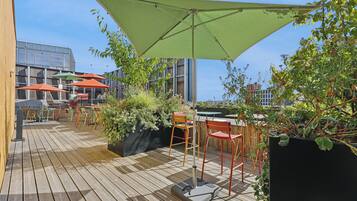 An outdoor patio area with wooden flooring, multiple tables and chairs, and a large green umbrella providing shade.