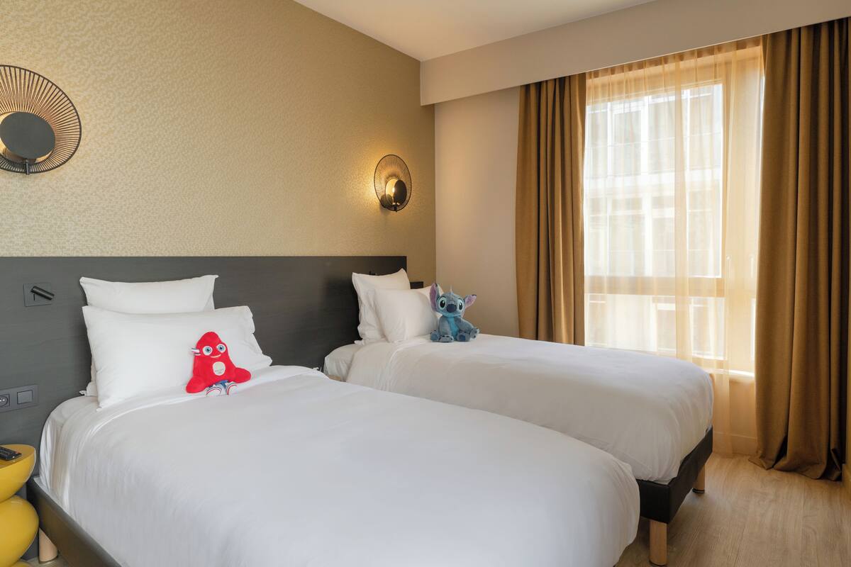 A hotel room with two beds, a wall-mounted light fixture, and a large window with curtains.