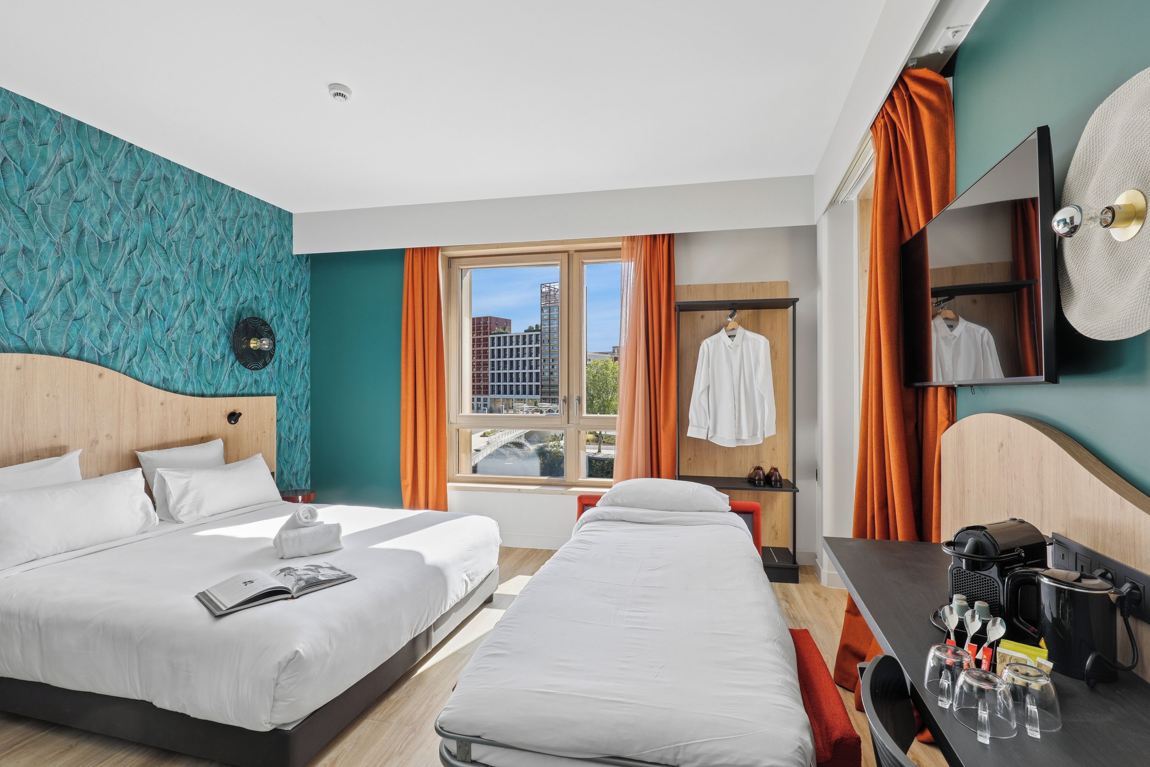 A hotel room with two beds, a desk, a mirror, and a window with a view of buildings.