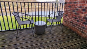 A balcony with two wicker chairs and a small table, overlooking a grassy area and a body of water.