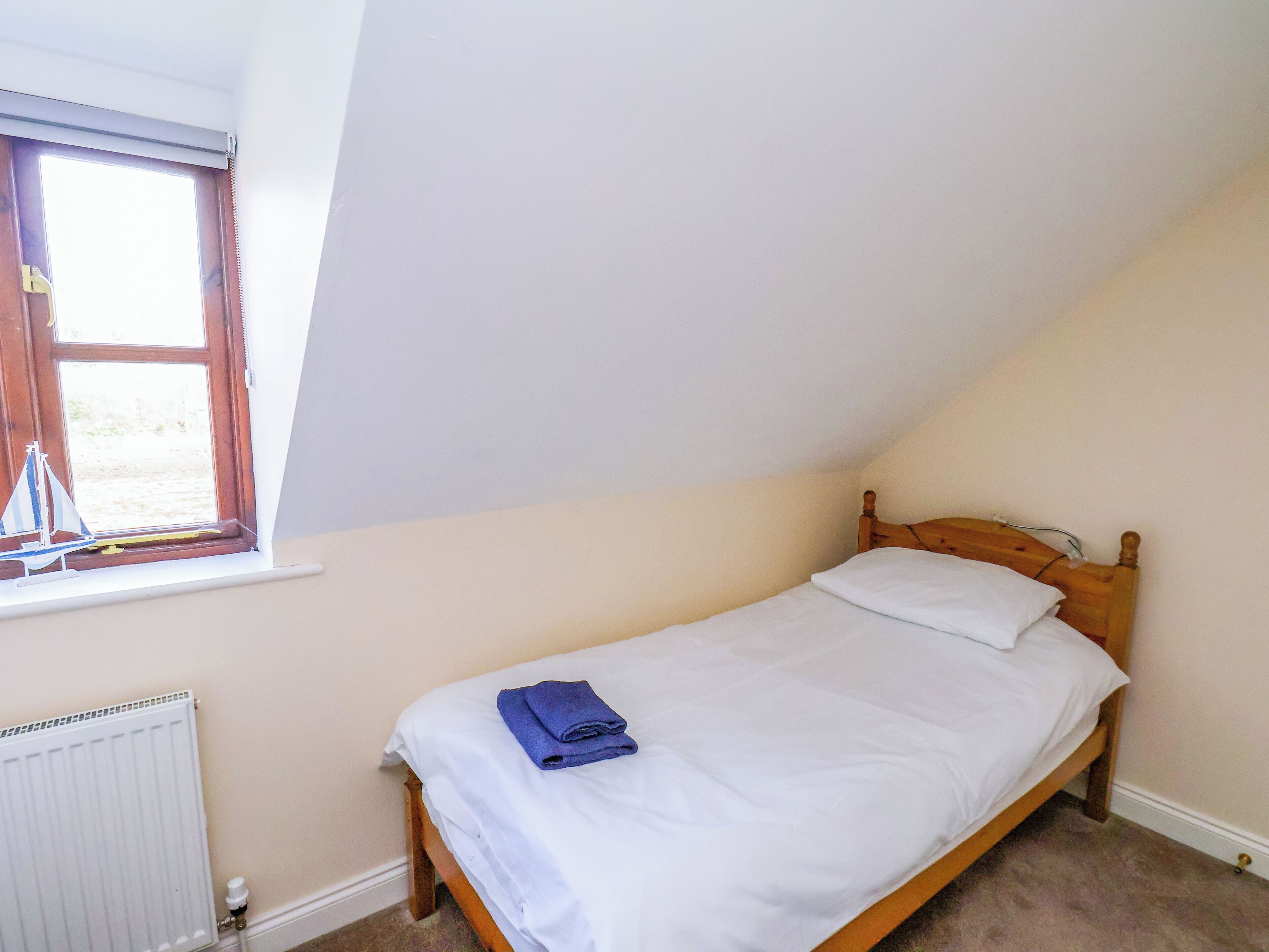 Ein kleines Zimmer mit einem Einzelbett, geneigter Decke, einem Fenster mit Jalousien, einem Heizkörper und einem Holzkopfende.