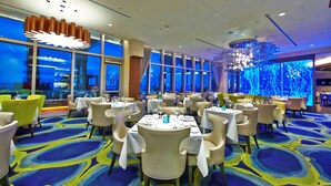 Ein Restaurant mit runden Tischen und Stühlen, großen Fenstern und einem blauen Teppich.
