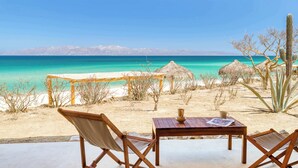 A beachside seating area with a table and chairs, overlooking a turquoise sea and sandy beach.