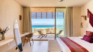 A hotel room with a bed, a desk, a chair, a surfboard, and a view of the beach.