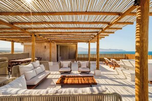 Un área de estar al aire libre con una pérgola de madera, asientos acolchados y vistas al mar.
