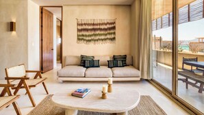 A modern living room with a beige sofa, two wooden chairs, a round coffee table with candles, and a wall hanging.