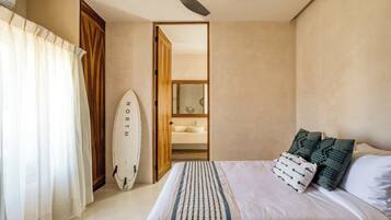 A bedroom with a bed, a ceiling fan, a surfboard, and a bathroom visible through an open door.