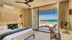 Habitación de hotel con cama, una banca, ventilador de techo y balcón con vista a la playa y al océano.