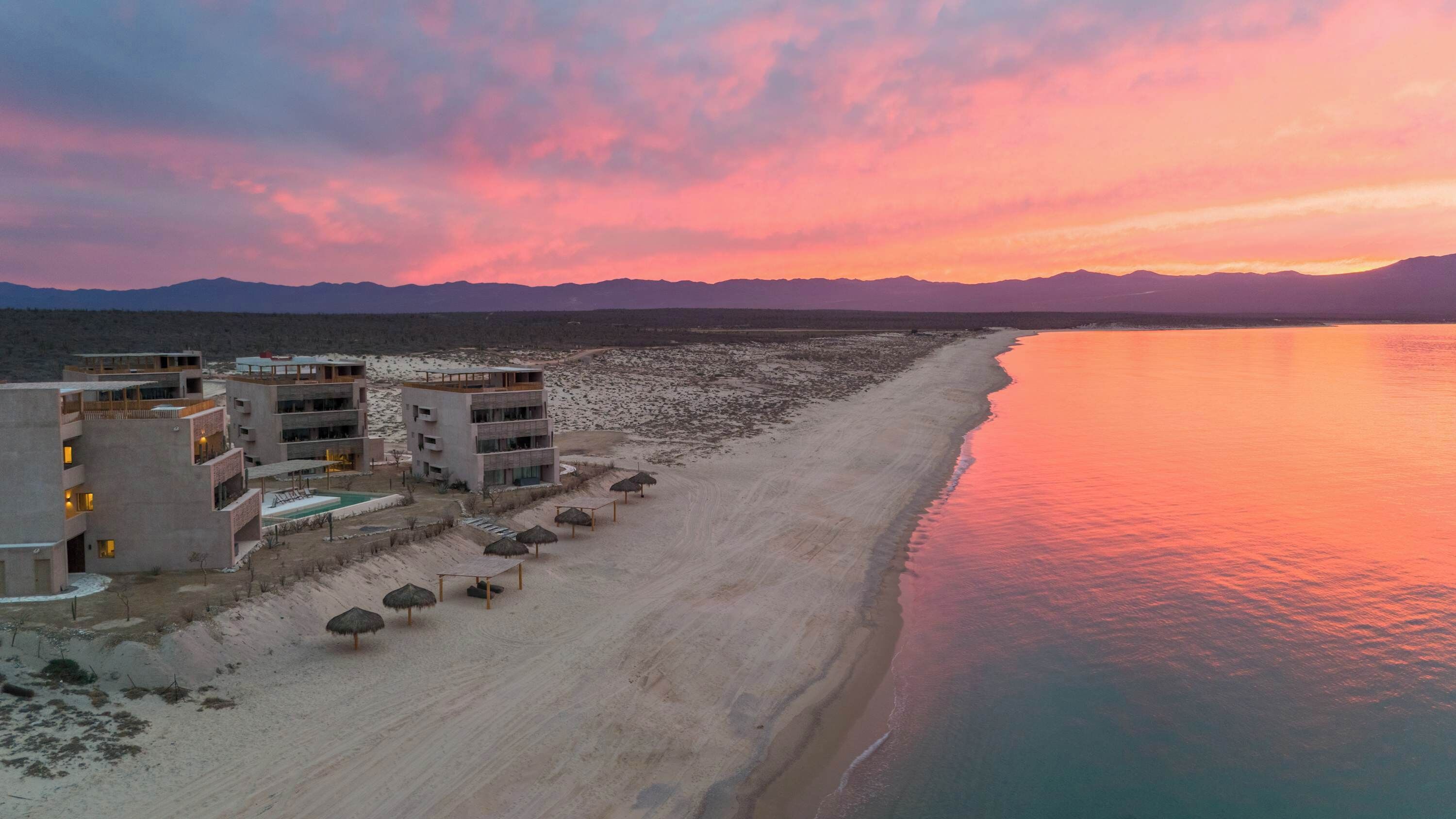 A coastal hotel with multiple floors, a swimming pool, and beach umbrellas.