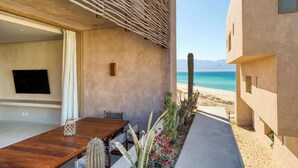 A modern outdoor dining area with a wooden table and chairs, a cactus, and a view of the beach and mountains. - Oceanview Vista Villa (La Ventana)