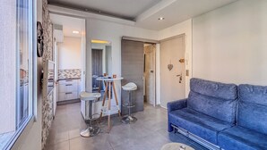 A modern living room with a blue sofa, a small round table with a vase, and a kitchen area with a dining table and chairs. - Contemporary 2P Studio Just Steps from the Beach (Cagnes-sur-Mer)