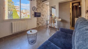 A modern living room with a large window, a stone wall, a TV, and a seating area. - Contemporary 2P Studio Just Steps from the Beach (Cagnes-sur-Mer)