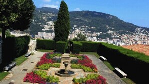 A garden pathway with a fountain, benches, and colorful flowers leading to a cityscape with mountains in the background. - Apartment for 8 guests in Nice city center (Nice)