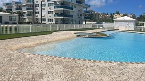 A swimming pool with a tiled deck, surrounded by a white fence. In the background, there is a multi-story building with balconies and a clear blue sky. - Cozy Studio in CostaMar Beach Village PR (Medianía Alta)