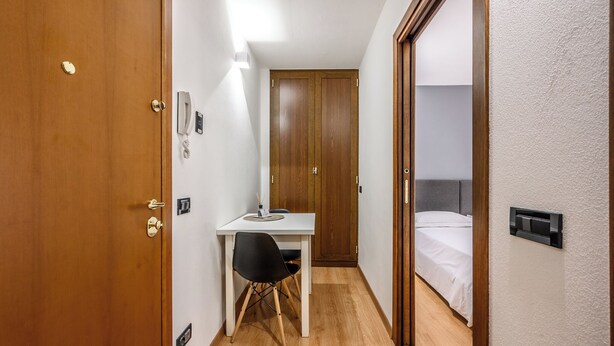 Ett hotellrum med en dörr av trä, ett skrivbord med en stol, en säng och ett litet bord med en lampa.
