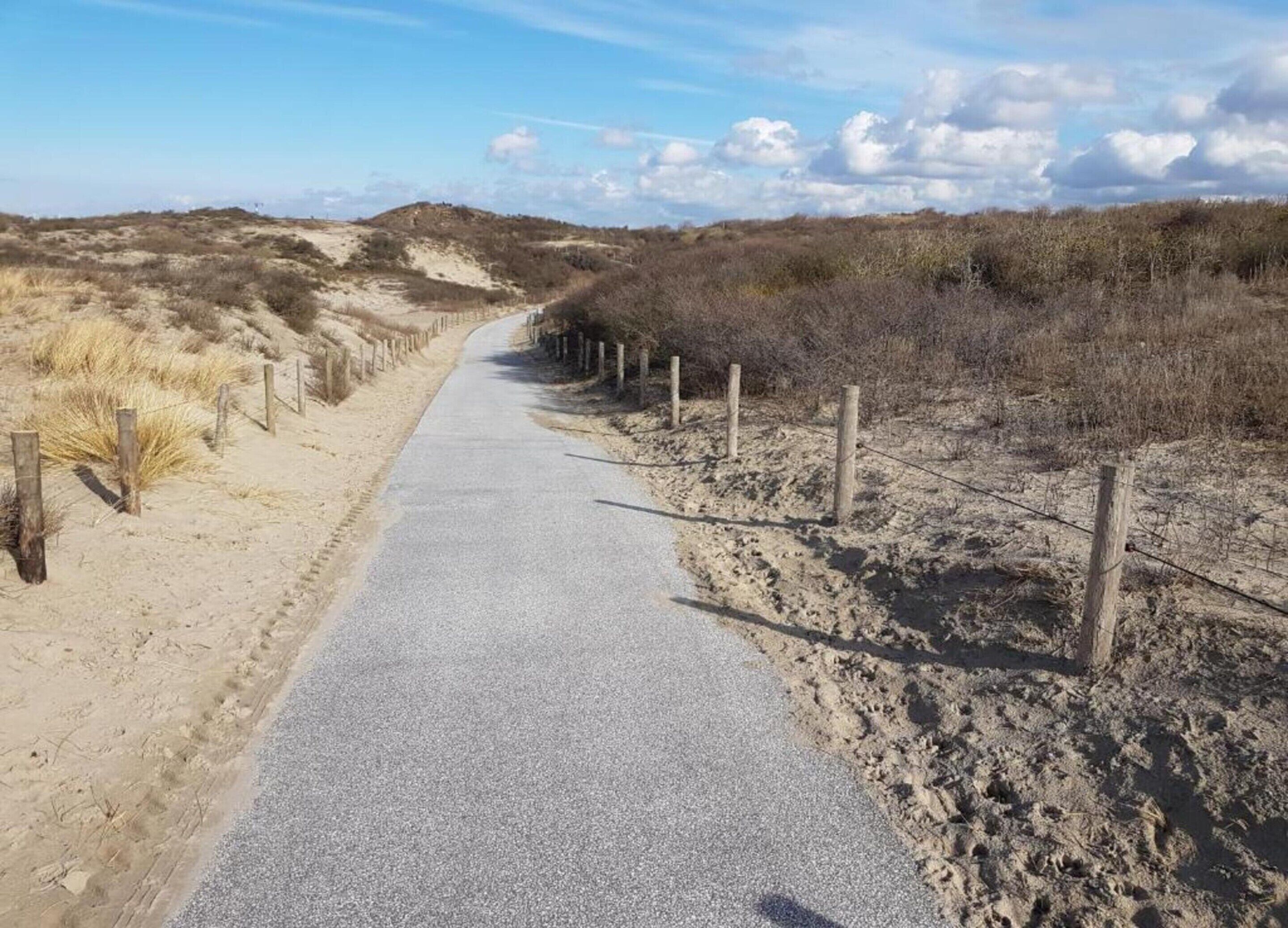 A paved pathway flanked by wooden fences and sparse vegetation, leading through a dry, hilly landscape.
