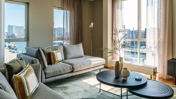 Un salon moderne avec un canapé gris, des coussins à motifs, une table basse ronde noire et une vue sur la ville grâce à de grandes fenêtres.