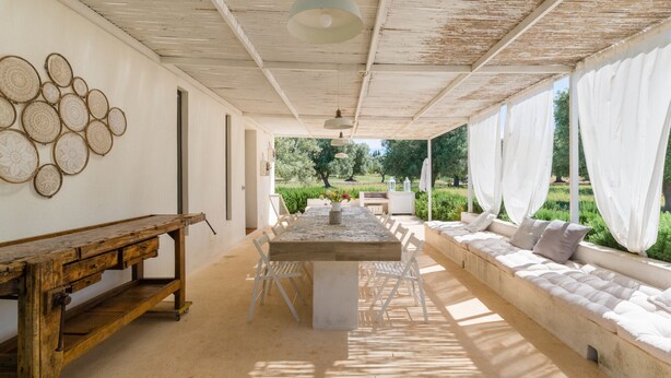 A covered outdoor dining area with a table and chairs, a wooden bench, and a sofa with cushions.
