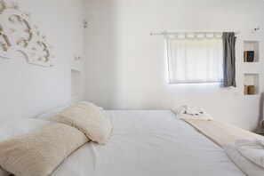 Un dormitorio con cama, almohadas, una ventana con cortinas y un aplique de pared.