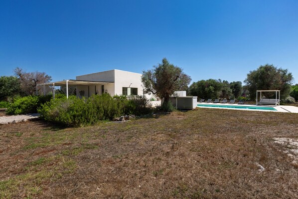 Una casa moderna con piscina, rodeada por un área cubierta de pasto seco y árboles.