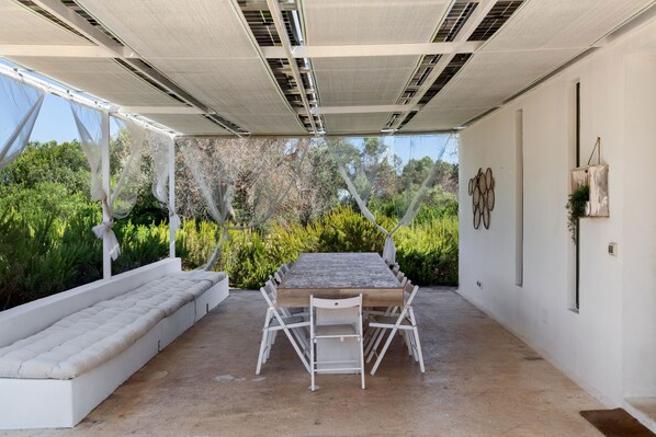 An outdoor patio with a table and chairs, a cushioned bench, and a view of greenery.